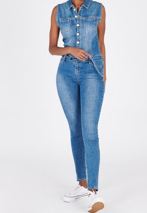 Kobieta ubrana w bezrękawnik z denimu zapinany na guziki oraz wysokie, obcisłe jeansy ze białymi trampkami, trzymająca pętlę paska z denimu.
