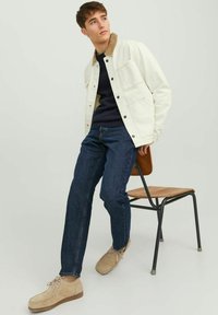 Bílá džínová bunda s hnědým manšestrovým límcem, navy svetr, tmavě modré džíny a béžové suede boty. Model sedí na dřevěné židli.