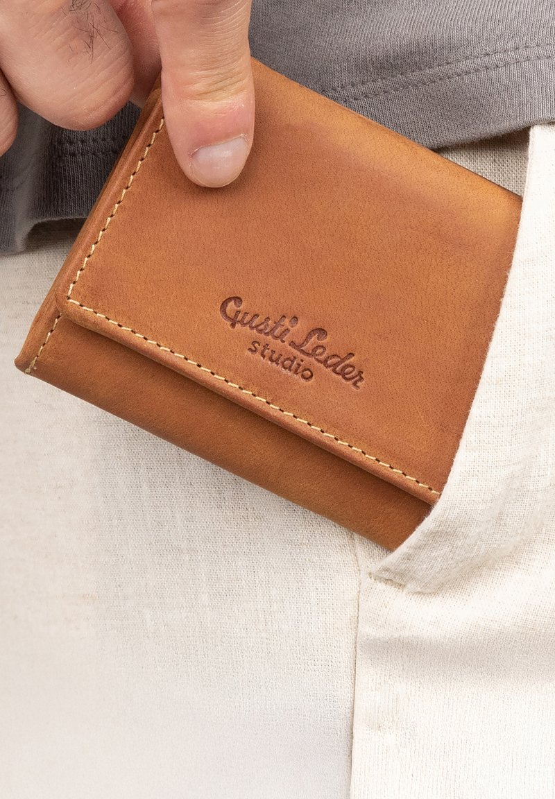 Mano che tiene un portafoglio in pelle beige con la scritta "Gusti Leder studio" impressa, parzialmente inserito in una tasca di pantaloni chiari.