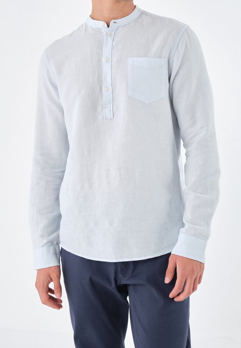 Man in een lichtblauwe lange mouwen henley shirt met borstzak en marineblauwe broek, staand tegen een witte achtergrond.