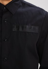 Närbild av en svart skjorta med knappar och krage, med en horisontell tygremsa ovanför det vänstra bröstfickområdet.