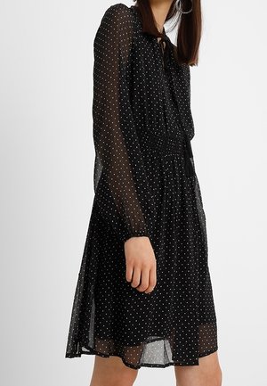 Femme portant une robe noire à manches longues avec des pois blancs et une taille cintrée, debout devant un fond blanc uni.