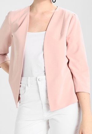 Femme portant un blazer rose clair ouvert sur un haut blanc et un pantalon blanc, debout avec une main sur la hanche devant un fond uni.