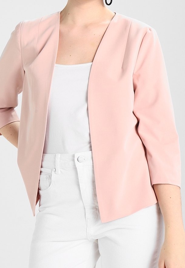 Femme portant un blazer rose clair ouvert sur un haut blanc et un pantalon blanc, debout avec une main sur la hanche devant un fond uni.