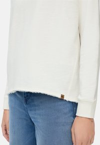 Witte lange mouwen shirt met een ruwe zoom, gedragen met lichtblauwe jeans. Aan de rechterzijde van de onderzoom van het shirt is een klein bruin label zichtbaar.