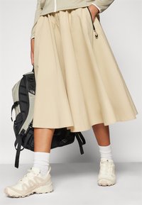 Beige, térdközépig érő szoknya, redőkkel és oldalsó zsebekkel, világos színű tornacipővel és fehér zoknival párosítva. Fekete hátizsák látható.