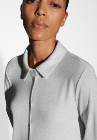Polo shirt a coste di colore grigio chiaro, con colletto e bottoni; il tessuto ha una leggera lucentezza e una texture liscia.