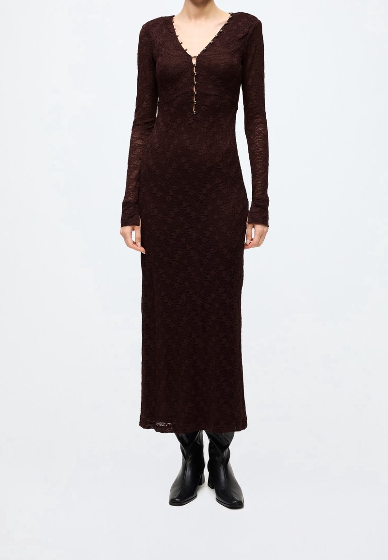 Robe longue en dentelle marron foncé avec manches longues et boutons sur le devant, portée avec des bottes en cuir noires mi-mollet sur un fond uni.