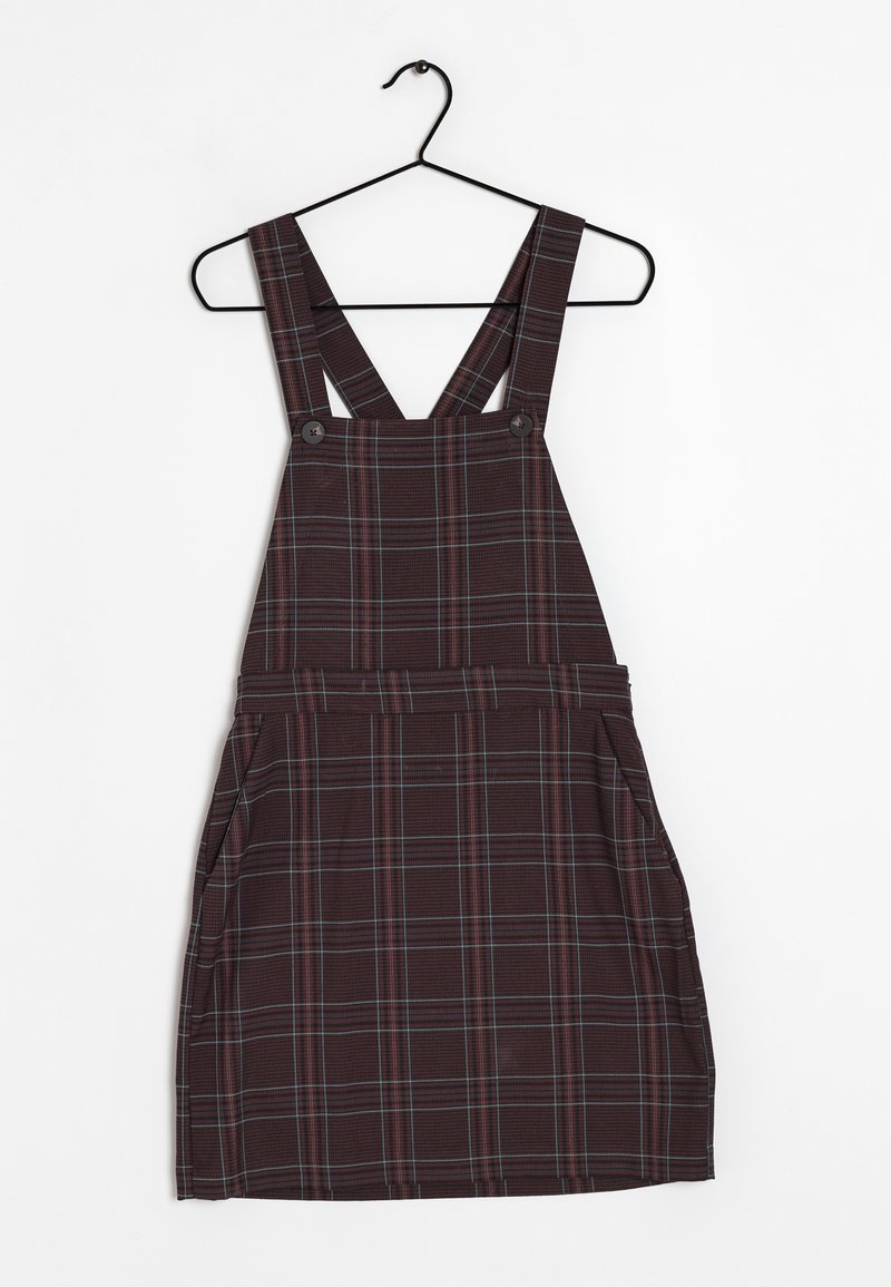 Robe tablier à carreaux foncés avec bretelles croisées et boutons, suspendue sur un cintre métallique noir sur fond blanc.
