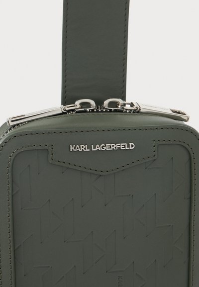 Крупен план на зелена чанта Karl Lagerfeld с изпъкнал логотип, видим шев и два сребърни ципа отгоре.