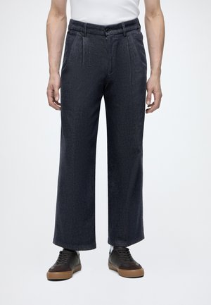 Mann trägt dunkle, weite Hosen, ein weißes Hemd, das in die Hose gesteckt ist, und schwarze Sneakers mit braunen Sohlen, steht vor einem einfarbigen hellen Hintergrund.
