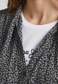 Camisa negra con estampado floral y pequeños patrones en azul y blanco, con cuello abotonado, superpuesta sobre una camiseta blanca con texto negro.