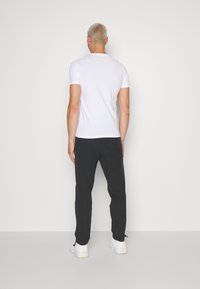 Weiße kurzärmlige T-Shirt und schwarze Hose, von hinten stehend. Die Hose hat elastische Bündchen und wird mit weißen Turnschuhen kombiniert.
