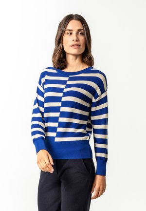 Frau trägt einen blau-weiß gestreiften Langarmpullover und schwarze Hose, steht mit einer Hand in der Nähe ihrer Taille und schaut nach rechts.