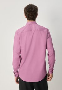 Langärmeliges Hemd aus einfarbigem pinkfarbenem Stoff, mit Knopfkragen, abgerundetem Saum und Knopfmanschetten. Glatte Textur ohne sichtbare Muster.