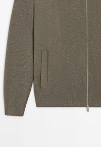 Olivgrüne Strickjacke mit durchgehendem Reißverschluss, Seitentasche und gerippten Bündchen sowie Saum. Weiche Textur mit dezentem Muster.