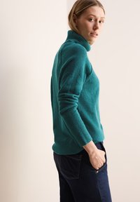 Türkiser Rollkragenpullover mit gerippten Ärmeln, tailliert geschnitten, kombiniert mit dunklen Jeans. Glatte Textur und minimales Design.