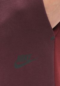 Maroonskrāsa sporta audums ar gludu tekstūru, ar melnu Nike logu un zīmi. Sānu šuvē norādīts ar mazu melnu akcentu.