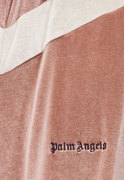 Ύφασμα βελούδου με μπεζ διαγώνια ρίγα και τη λέξη "Palm Angels" κεντημένη με μαύρη γοτθική γραμματοσειρά σε φόντο σκούρου ροζ.