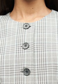 Κοντινό πλάνο ενός ατόμου που φοράει γκρι καρό ρούχο με τρία μεγάλα γκρι κουμπιά με την επιγραφή "GUESS" κατά μήκος του μπροστινού μέρους.