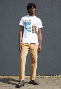 Vit t-shirt med grafiska tryck, beige byxor och bruna träskor. Modellen står mot en grå vägg och bär solglasögon.