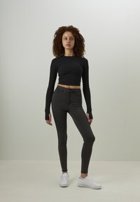 Svart langermet crop-top, grå høytlivs skinny jeans, og hvite sneakers; modell som står foran en nøytral bakgrunn med hendene ved sidene.