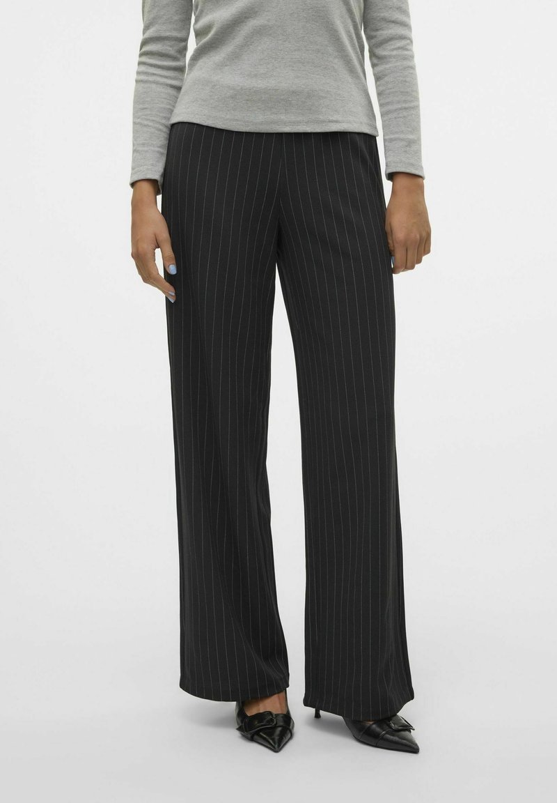 Femme portant un pantalon large noir à fines rayures et des talons pointus noirs, debout devant un fond clair uni.