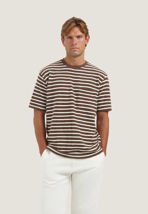 Uomo con capelli castano chiaro che indossa una camicia a maniche corte marrone e bianca a righe e pantaloni bianchi, in piedi con le mani nelle tasche davanti a uno sfondo neutro.