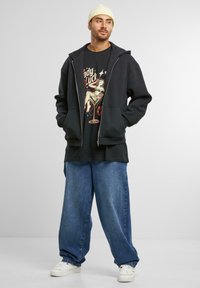 Hanorac negru cu fermoar, tricou grafic cu un design vintage, pantaloni denim bleumarin largi și adidași albi, completat cu o căciulă crem.