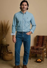 Man met halflang donker haar, gekleed in een lichtblauw overhemd, blauwe jeans en bruine schoenen, staande in een rustieke kamer met een stoel en een mand.