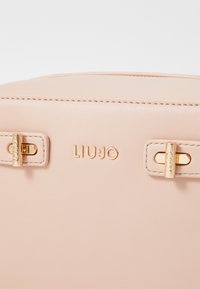 Hele roosa nahast käekott kuldsete detailidega, millel on sile tekstuur, brändi logo ja minimalistlikud aktsendid, et saavutada elegantne disain.