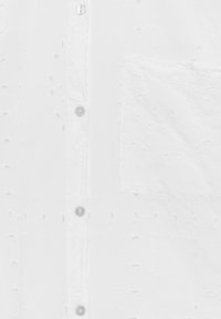 Chemise blanche boutonnée en tissu texturé avec détails brodés, quatre boutons et une poche plaquée sur le côté gauche.