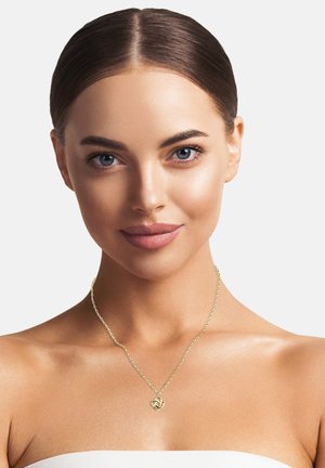 Collana d'oro con un piccolo pendente a nodo attorcigliato su una catena sottile, indossata su spalle scoperte. Il tono della pelle è liscio e chiaro.