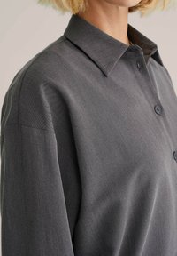 Närbild av en person som bär en mörkgrå skjorta med knappar och spetsig krage, med kort blont hår synligt.