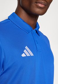 Μπλε πόλο μπλούζα από υφασμάτινο υλικό με γιακά, με λευκό λογ ότυπο adidas στο αριστερό στήθος.