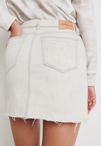 Bílá denimová mini sukně s třepeným lemem, má dvě zadní kapsy s viditelným šitím a kožený štítek s logem na pásku.