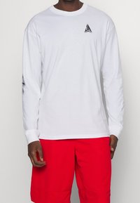 Weißes Langarmshirt mit einem schwarzen Logo auf der Brust und einem grafischen Muster an den Ärmeln. Getragen mit roten Shorts.