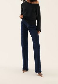 Femme portant un pull côtelé sombre à épaules dénudées, un jean bleu foncé taille haute à jambes droites, et des talons pointus couleur nude avec un détail nœud.