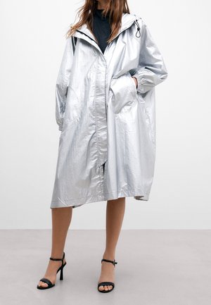 Frau trägt einen glänzenden silbernen knieumspielenden Kapuzenmantel mit Taschen, kombiniert mit schwarzen Sandalen mit Absatz und einem schwarzen Oberteil darunter.