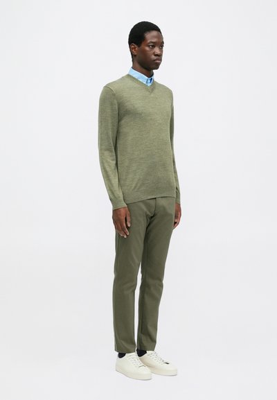 Uomo in piedi che indossa un maglione verde chiaro a V, una camicia blu con bottoni, pantaloni oliva, calzini neri e sneakers bianche su sfondo bianco.