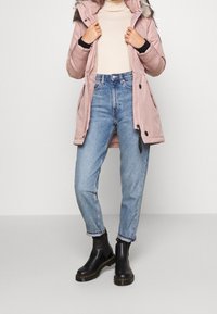 Ljusrosa parkas med huva i pälskant, svarta detaljer, som bärs med högmidjade blå jeans och svarta ankelboots. Slät textur genom hela plagget.