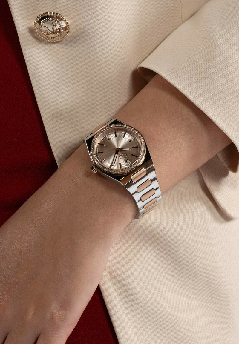 Ceas de mână argintiu și roz aurit cu un cadran texturat, bezel decorat cu pietre și o brățară metalică cu linkuri dreptunghiulare.