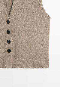 Gilet beige sans manches en tricot avec quatre boutons noirs sur le devant et bord côte sur fond blanc.