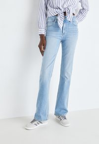 Lys blå bootcut jeans laget av denim, med en klassisk fem-lomme design og subtil slitasje, kombinert med grå sneakers og en stripete skjorte.