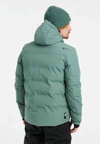 Grüne Puffjacke mit Kapuze, die ein gestepptes Muster, elastische Bündchen und Logo-Details aufweist. Trägt eine grüne Strickmütze.
