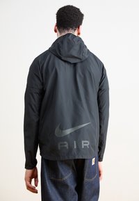 Černá větrovka s kapucí, je na ní reflexní logo Nike a nápis "AIR" na zádech. Materiál vypadá hladký a lehký.