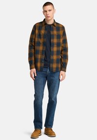 Bruine en marineblauwe geruite overhemd, met knopen aan de voorkant, lange mouwen, gedragen over een marineblauw T-shirt, gecombineerd met blauwe jeans en beige laarzen.