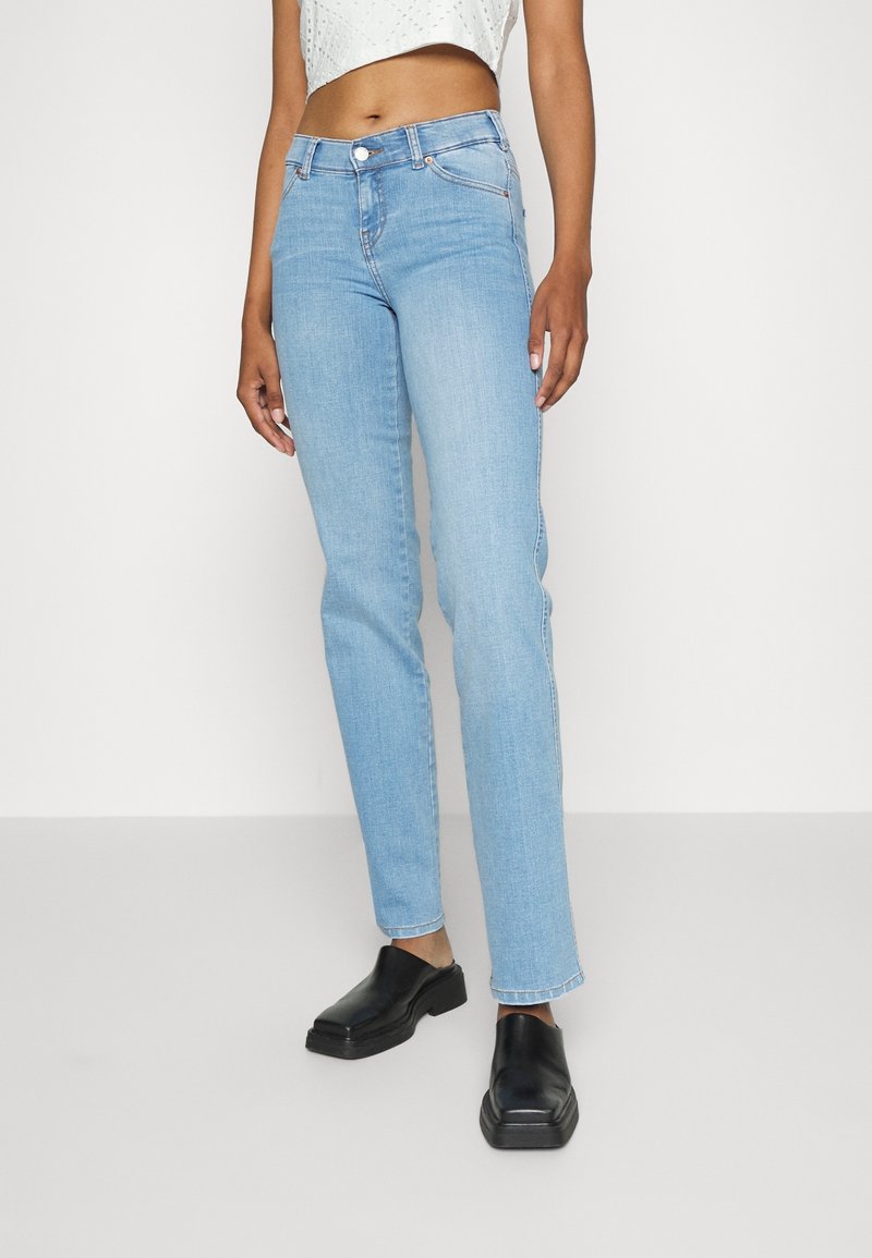 Jean en denim bleu clair avec une silhouette à jambes droites, un design à cinq poches et une taille haute. Porté avec des chaussures noires sans lacets.