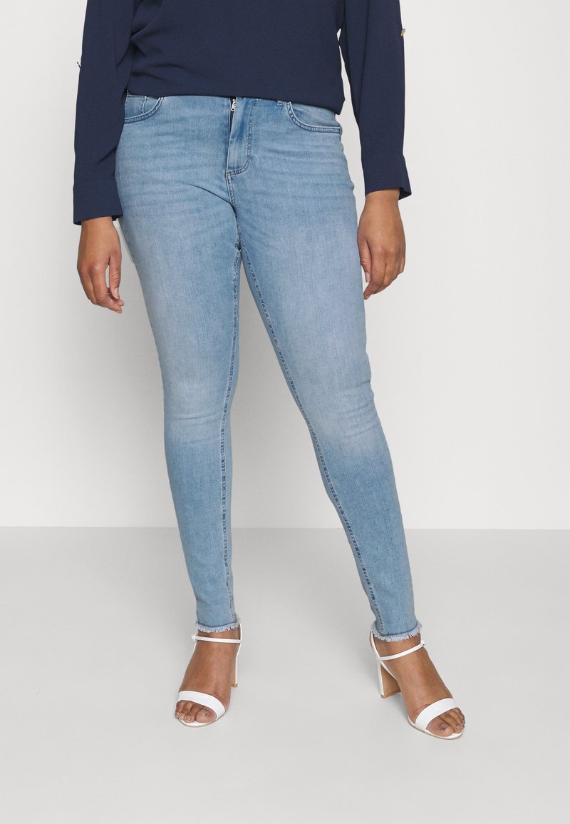 Světle modré skinny džíny vyrobené z denimu s vybledlou texturou, obsahující zipové zapínání, pět kapes a detaily s neupraveným lemem. Sestaveno s bílými lodičkami na podpatku.