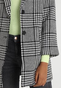 Blazer a motivi houndstooth bianco e nero con due bottoni, indossato sopra un top in maglia verde chiaro, abbinato a jeans neri slim-fit.